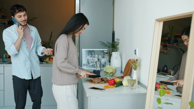 Mujer joven cocinando comida mientras su marido amado conversa por teléfono móvil y besando a su esposa en la cocina de su casa