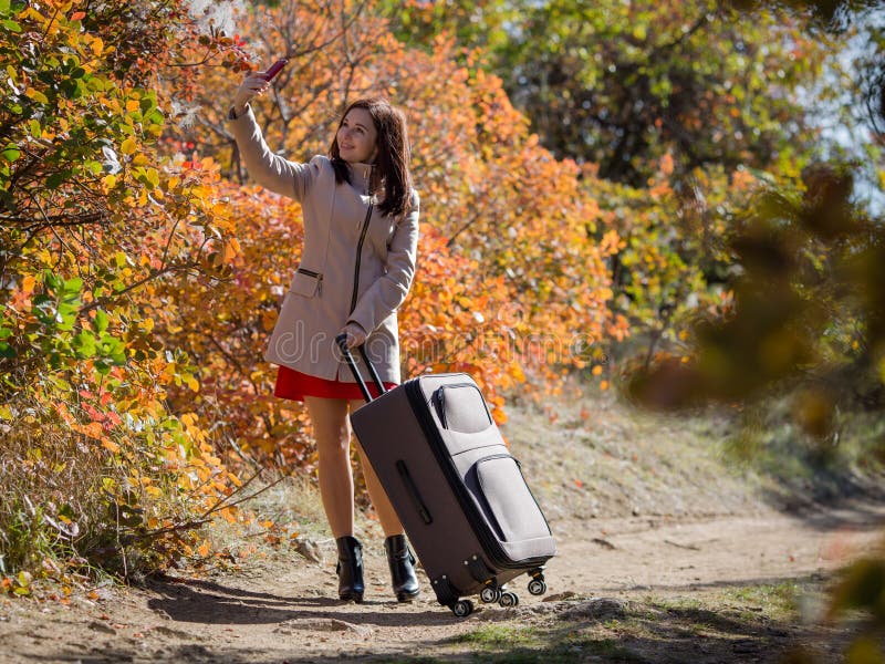 Mujer joven con el equipaje en la carretera nacional en la persona femenina del bosque en vestido rojo corto y la capa que toman foto de archivo libre de regalías Mujer joven con el equipaje en la carretera nacional en la persona femenina del bosque en vestido rojo corto y la capa que toman foto de archivo libre de regalías