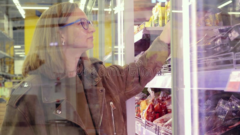 Mujer joven con gafas selecciona comidas congeladas de un congelador de supermercados. sujetando un contenedor de plástico negro almacen de video