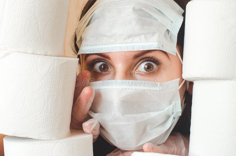 Mujer joven con grandes ojos asustados en dos caras de protección contra virus médicos mira a través de pilas de papel higiénico. imagen de archivo