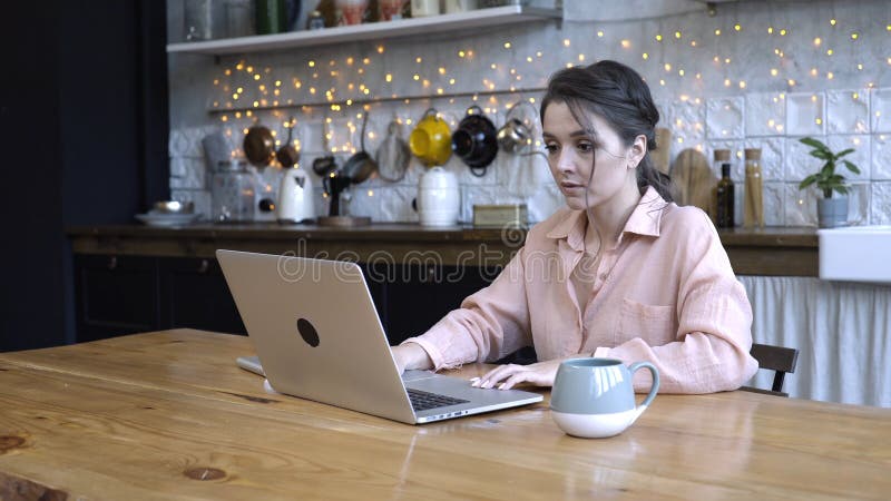 Mujer joven concentrada sentada en la cocina en una mesa de madera y tecleando en su portátil personal de plata con la taza de foto de archivo Mujer joven concentrada sentada en la cocina en una mesa de madera y tecleando en su portátil personal de plata con la taza de foto de archivo