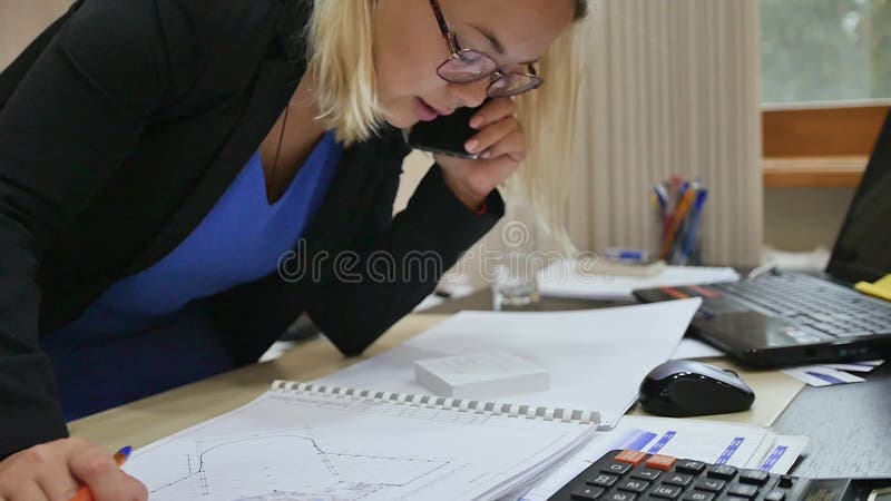 Mujer joven de negocios habla por teléfono en la oficina, examinando los planos en la mesa