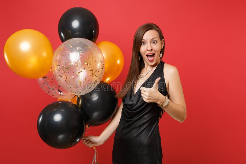 Mujer joven emocionada en poco vestido negro que celebra mostrando el pulgar encima de sostener los balones de aire aislados en r foto de archivo libre de regalías Mujer joven emocionada en poco vestido negro que celebra mostrando el pulgar encima de sostener los balones de aire aislados en r foto de archivo libre de regalías