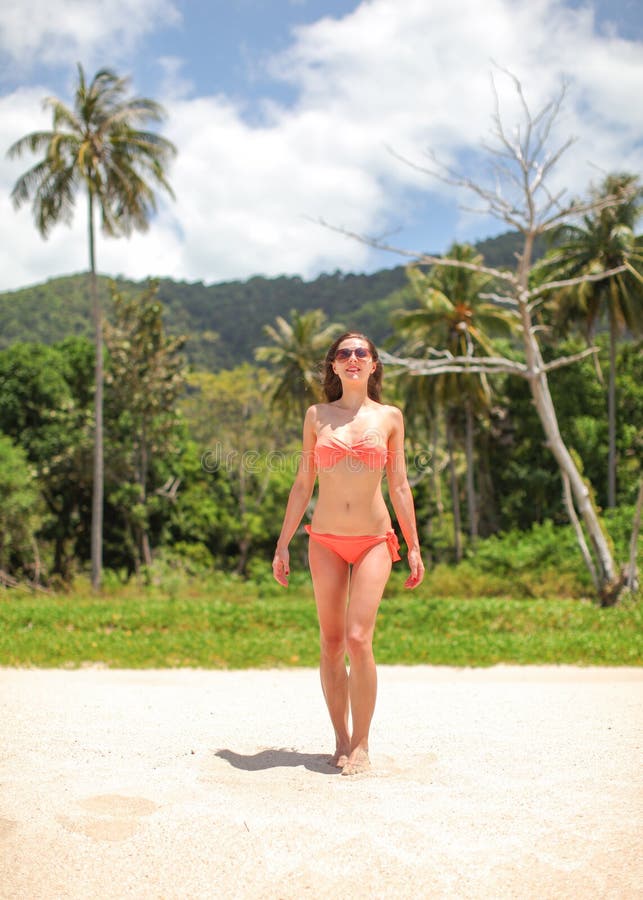 Mujer joven en bikini y gafas de sol rojos que camina en la playa, selva con las palmeras detrás de ella foto de archivo libre de regalías Mujer joven en bikini y gafas de sol rojos que camina en la playa, selva con las palmeras detrás de ella foto de archivo libre de regalías