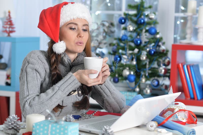 Mujer joven en sombrero de Santa con taza de té sentada frente a una laptop fotos de archivo libres de regalías Mujer joven en sombrero de Santa con taza de té sentada frente a una laptop fotos de archivo libres de regalías