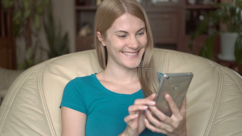 Mujer joven feliz alegre que se sienta en silla en casa Usando su smartphone, hojeando, leyendo noticias almacen de metraje de vídeo