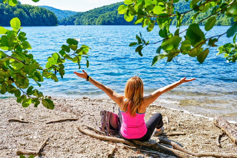 Mujer joven feliz con sus brazos separados en la alegría, sentándose en una orilla del lago de la montaña, llevando una camisa fu fotos de archivo libres de regalías Mujer joven feliz con sus brazos separados en la alegría, sentándose en una orilla del lago de la montaña, llevando una camisa fu fotos de archivo libres de regalías