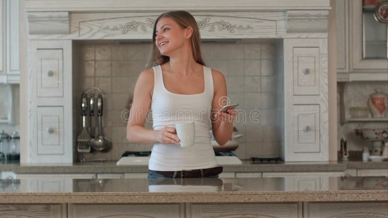 Mujer joven feliz hermosa con café de consumición del teléfono en casa foto de archivo Mujer joven feliz hermosa con café de consumición del teléfono en casa foto de archivo