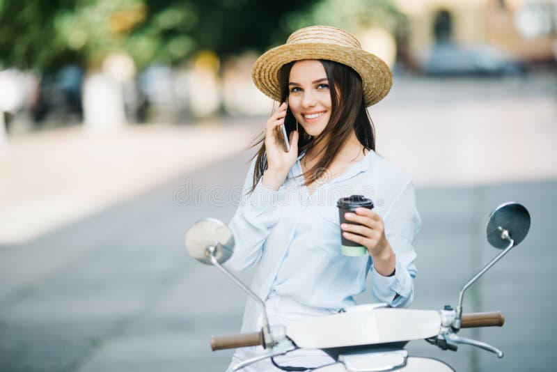 Mujer joven feliz que bebe el café para llevar cerca de su ciclomotor y que habla el teléfono en la calle de la ciudad imagen de archivo libre de regalías Mujer joven feliz que bebe el café para llevar cerca de su ciclomotor y que habla el teléfono en la calle de la ciudad imagen de archivo libre de regalías