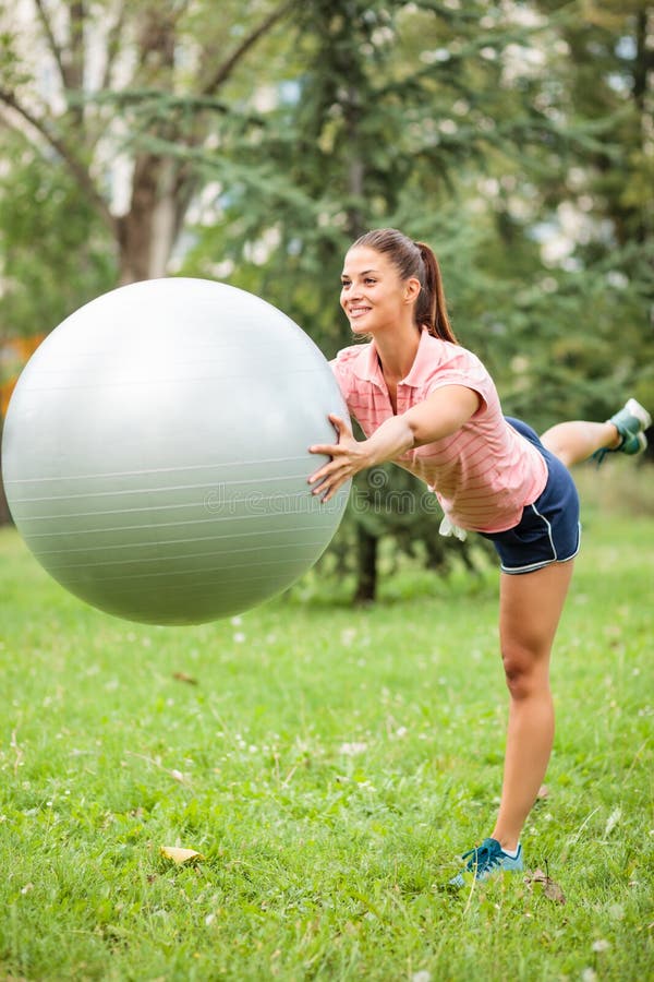Mujer joven feliz que equilibra en una pierna y que sostiene la bola de la aptitud delante de su pecho fotografía de archivo libre de regalías Mujer joven feliz que equilibra en una pierna y que sostiene la bola de la aptitud delante de su pecho fotografía de archivo libre de regalías