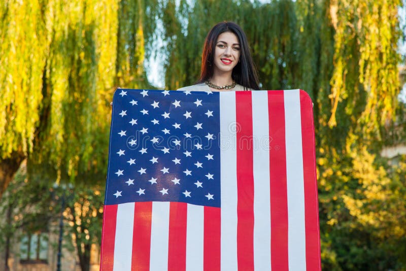 Mujer joven hermosa con el vestido clásico que sostiene la bandera americana en el parque modelo de moda que nos detiene que sonr imagen de archivo libre de regalías Mujer joven hermosa con el vestido clásico que sostiene la bandera americana en el parque modelo de moda que nos detiene que sonr imagen de archivo libre de regalías