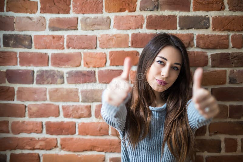 Mujer joven hermosa en ropa de sport con los pulgares para arriba, sonrisa, oponiéndose a la pared de ladrillo blanca fotos de archivo libres de regalías Mujer joven hermosa en ropa de sport con los pulgares para arriba, sonrisa, oponiéndose a la pared de ladrillo blanca fotos de archivo libres de regalías