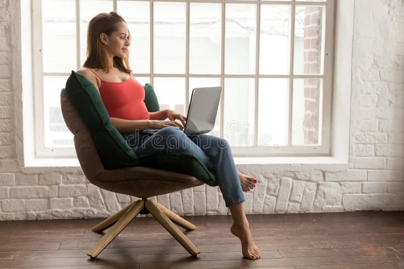 Mujer joven hermosa que se sienta en silla cómoda y que usa el ordenador portátil imagen de archivo Mujer joven hermosa que se sienta en silla cómoda y que usa el ordenador portátil imagen de archivo