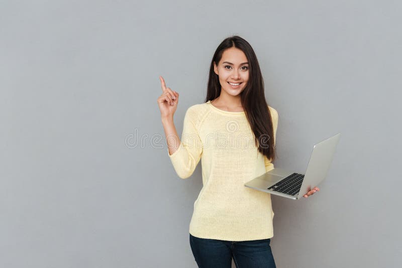 Mujer joven hermosa sonriente que sostiene el ordenador portátil y que señala lejos imágenes de archivo libres de regalías Mujer joven hermosa sonriente que sostiene el ordenador portátil y que señala lejos imágenes de archivo libres de regalías