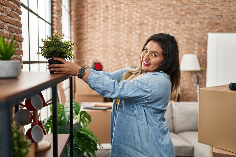 Mujer joven, hermosa y grande, sonriente y segura de poner la planta en el estante en su nuevo hogar imagen de archivo libre de regalías