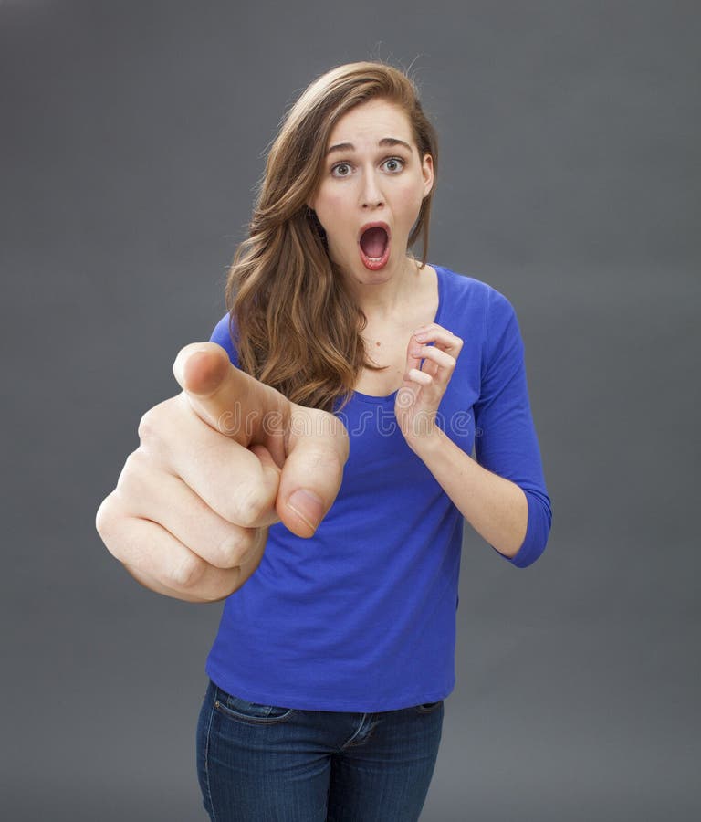 Stunned beautiful young woman with surprising mouth opened, pointing at someone with a large index for concept of danger, risk, accusation or recognition, grey background. Stunned beautiful young woman with surprising mouth opened, pointing at someone with a large index for concept of danger, risk, accusation or recognition, grey background