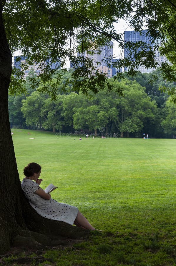 Mujer joven que lee un libro debajo de un árbol en el Central Park con el horizonte de Nueva York en el fondo, en la ciudad de Nu imágenes de archivo libres de regalías Mujer joven que lee un libro debajo de un árbol en el Central Park con el horizonte de Nueva York en el fondo, en la ciudad de Nu imágenes de archivo libres de regalías