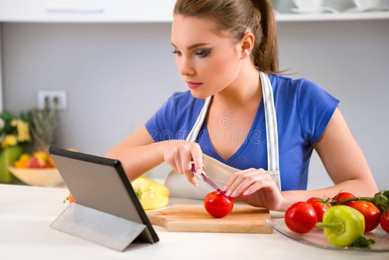Mujer joven que prepara la comida y que mira la tableta fotos de archivo Mujer joven que prepara la comida y que mira la tableta fotos de archivo