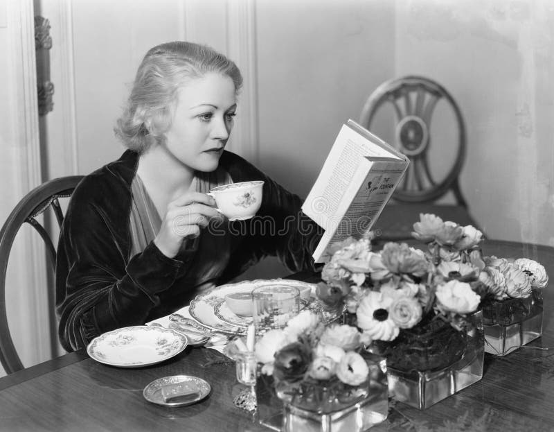 Mujer joven que se sienta en la mesa de comedor que sostiene una taza de té y que lee un libro (todas las personas representadas fotos de archivo Mujer joven que se sienta en la mesa de comedor que sostiene una taza de té y que lee un libro (todas las personas representadas fotos de archivo