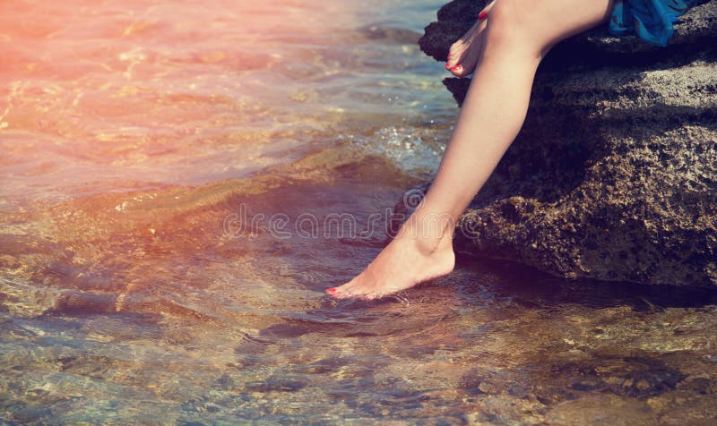Mujer joven que se sienta en una piedra, caída pies en la agua de mar imagen de archivo Mujer joven que se sienta en una piedra, caída pies en la agua de mar imagen de archivo