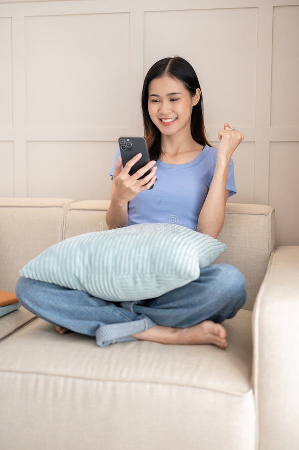 Mujer joven que usa smartphone y celebra sus buenas noticias sentada en el sofá imagen de archivo libre de regalías