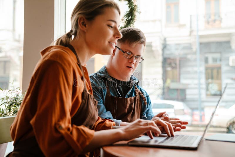 Mujer joven que usa una laptop mientras entrena a un hombre con síndrome de baja para trabajar en una cafetería fotos de archivo