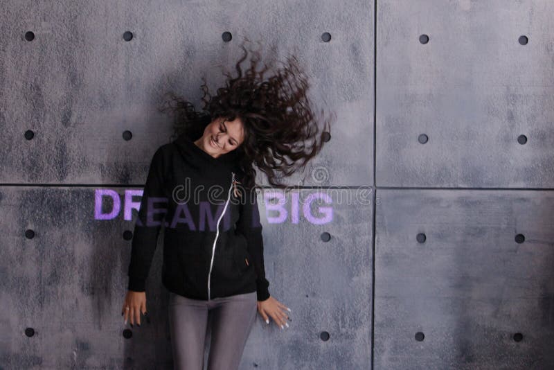 Mujer joven rizada con el pelo volador posando activamente en el fondo de la pared de concreto. Un sueÃ±o grande iluminado de colo imagen de archivo