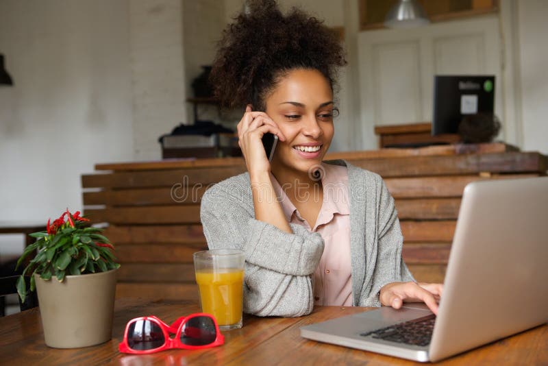 Mujer joven sonriente que usa el ordenador portátil y hablando en el teléfono móvil fotografía de archivo Mujer joven sonriente que usa el ordenador portátil y hablando en el teléfono móvil fotografía de archivo