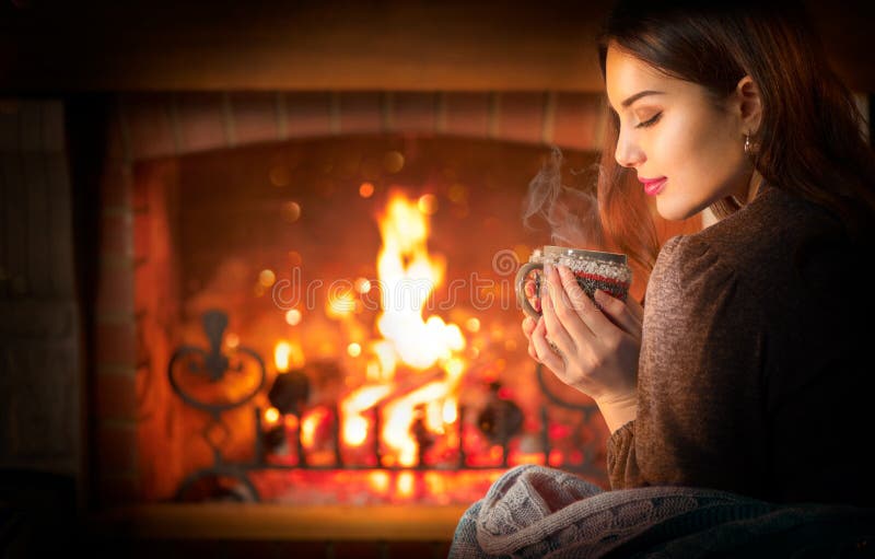 Mujer joven y bella de Navidad sentada cerca de la chimenea en una habitación oscura en casa y tomando bebidas calientes de una t imagen de archivo libre de regalías