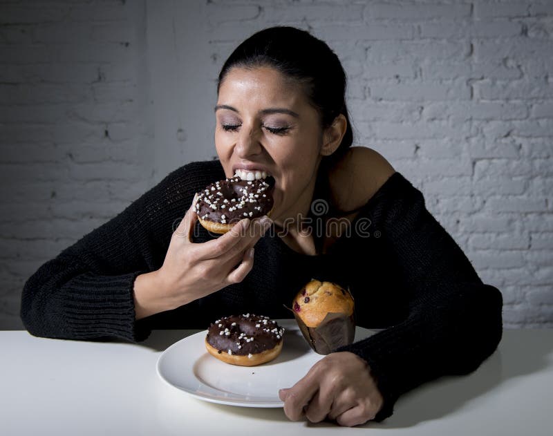 Young attractive latin woman sitting at table eating dish full of junk sugary unhealthy food such as chocolate donut and muffin cake in sugar addiction and hell with diet concept. Young attractive latin woman sitting at table eating dish full of junk sugary unhealthy food such as chocolate donut and muffin cake in sugar addiction and hell with diet concept