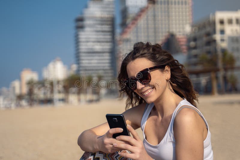 Mujer linda joven que usa smartphone en la playa Horizonte de la ciudad en fondo fotos de archivo Mujer linda joven que usa smartphone en la playa Horizonte de la ciudad en fondo fotos de archivo