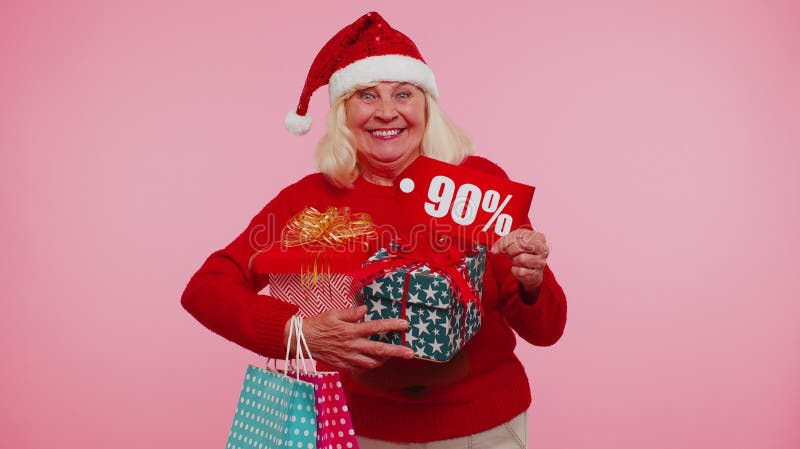 Senior grandmother woman in Christmas sweater showing gift box and 90 Percent discount inscriptions banner text. Black Friday. Good holiday low prices for shopping. Blue studio background. New Year. Senior grandmother woman in Christmas sweater showing gift box and 90 Percent discount inscriptions banner text. Black Friday. Good holiday low prices for shopping. Blue studio background. New Year