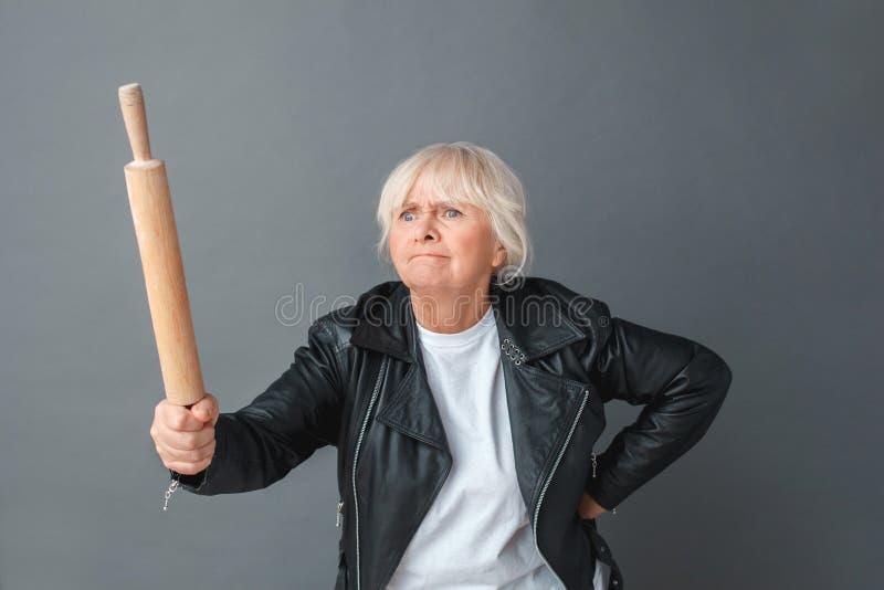 Mujer mayor en la situación del estudio de la chaqueta de cuero aislada en gris con el rodillo que amenaza alguien enojado fotografía de archivo Mujer mayor en la situación del estudio de la chaqueta de cuero aislada en gris con el rodillo que amenaza alguien enojado fotografía de archivo