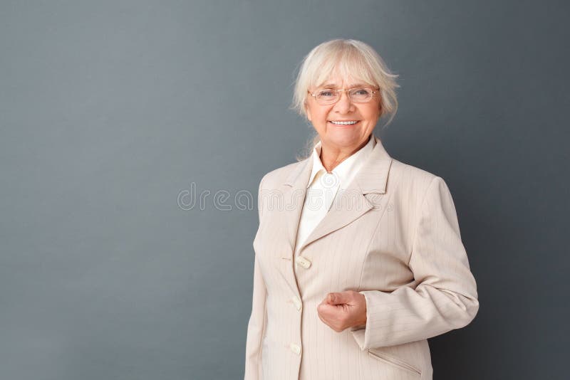 Mujer mayor en traje fromal y los vidrios que colocan el estudio en la cámara de mirada de presentación gris alegre imagen de archivo libre de regalías Mujer mayor en traje fromal y los vidrios que colocan el estudio en la cámara de mirada de presentación gris alegre imagen de archivo libre de regalías