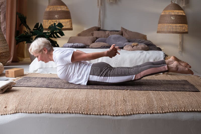 Mujer mayor practicando yoga en casa, haciendo pose de salabhasana o langosta