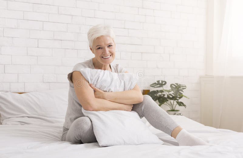 Happy senior woman resting on bed, hugging white pillow at home, lifestyle portrait. Happy senior woman resting on bed, hugging white pillow at home, lifestyle portrait