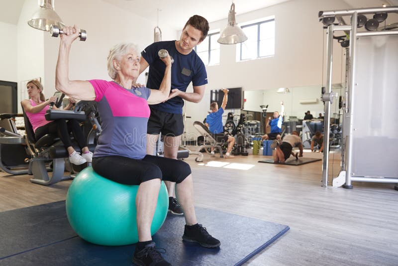 Mujer mayor que ejercita en bola suiza con los pesos que son animados por el instructor personal In Gym fotos de archivo Mujer mayor que ejercita en bola suiza con los pesos que son animados por el instructor personal In Gym fotos de archivo