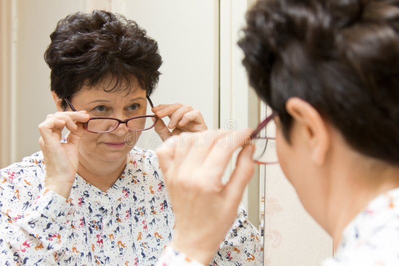 Mujer mayor que intenta en las nuevas lentes y que mira sobre los vidrios en el espejo imágenes de archivo libres de regalías Mujer mayor que intenta en las nuevas lentes y que mira sobre los vidrios en el espejo imágenes de archivo libres de regalías