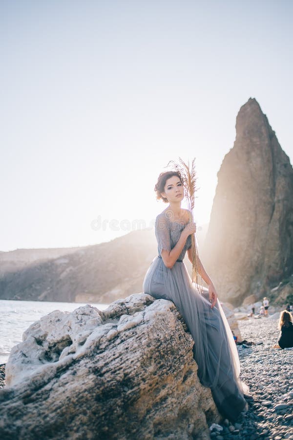Mujer morena hermosa en piedra blanca que se sienta del vestido largo el mar en la puesta del sol y mirando la cámara foto de archivo Mujer morena hermosa en piedra blanca que se sienta del vestido largo el mar en la puesta del sol y mirando la cámara foto de archivo