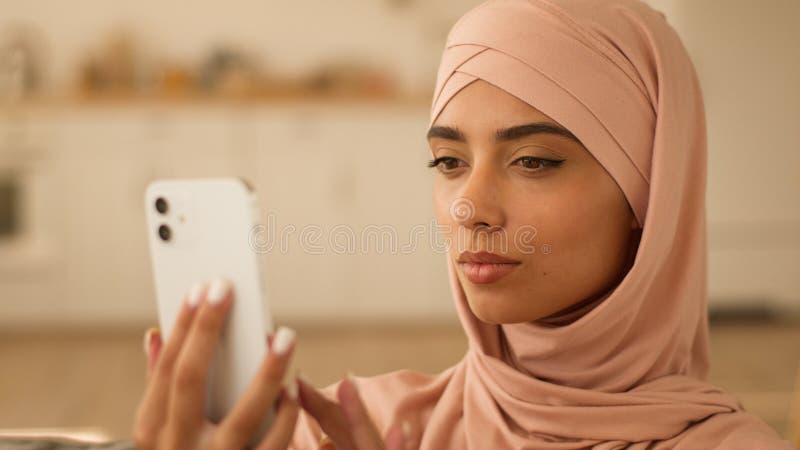 Mujer musulmana grave Niña islámica de etnia hembra con hijab en casa usando teléfonos móviles escribiendo un smartphone foto de archivo