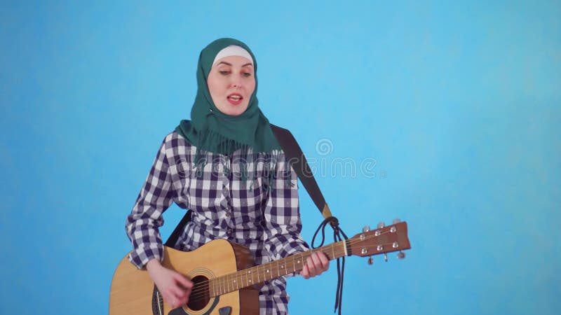 Mujer musulmán joven alegre que toca expresivo la guitarra en un fondo azul