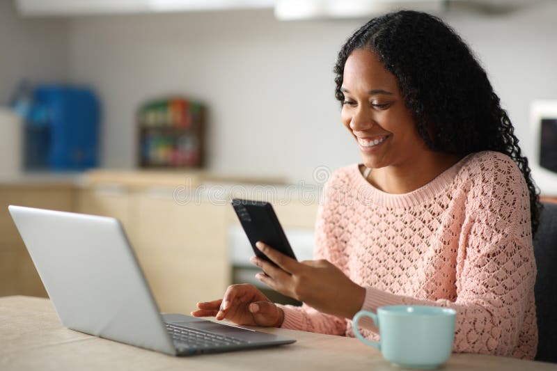 Mujer negra feliz usando teléfono y laptop en la cocina fotos de archivo