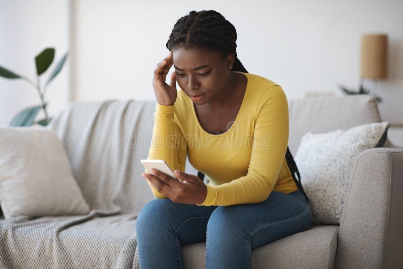 Mujer negra preocupada mirando la pantalla del smartphone, sentada en el sofá en casa