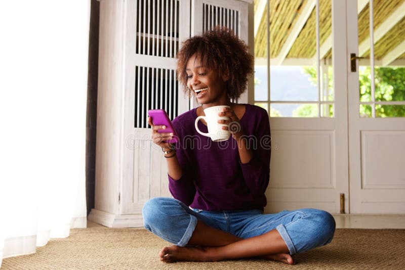Mujer negra sonriente que se sienta en piso en casa con el teléfono celular imagenes de archivo