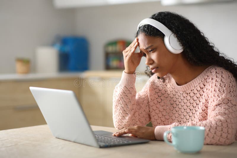 Mujer negra triste que se queja de comprobar el portátil en una cocina. Mujer negra triste usando auriculares quejándose de comprobar su laptop en una cocina en casa
