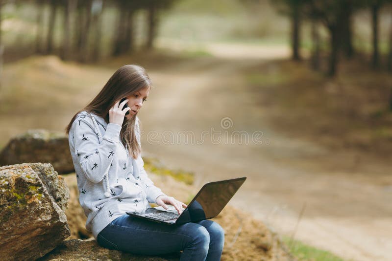 Mujer o estudiante en cuestión joven de negocios en la ropa casual que se sienta en piedra que habla en el teléfono móvil en parq fotografía de archivo Mujer o estudiante en cuestión joven de negocios en la ropa casual que se sienta en piedra que habla en el teléfono móvil en parq fotografía de archivo