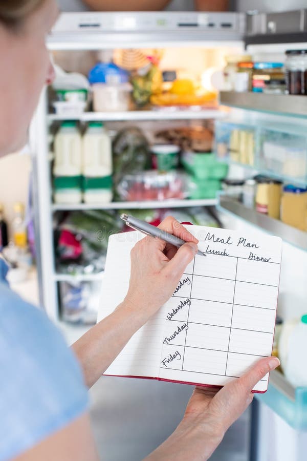 Mujer parada frente al refrigerador en la cocina con cuaderno escribiendo un plan semanal de comidas