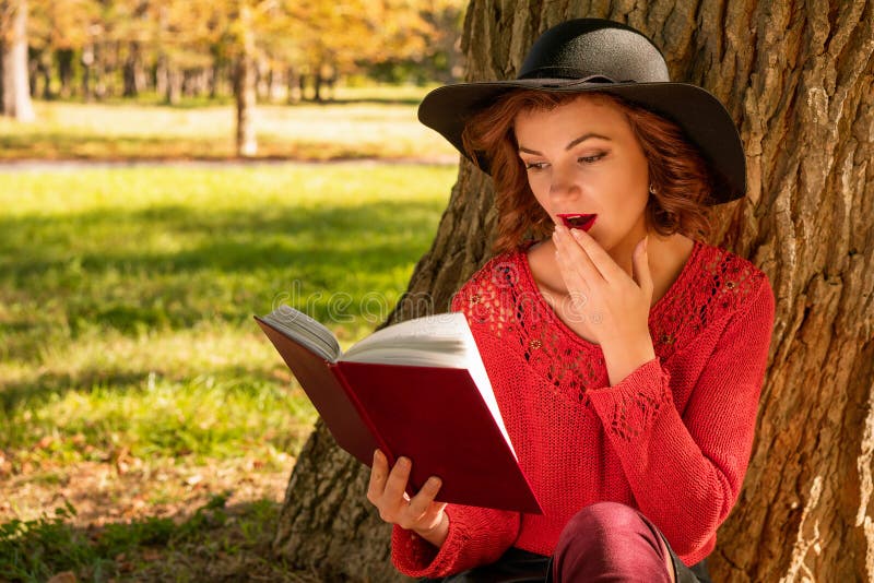 Mujer preciosa que lee un libro en el parque del otoño que se sienta en la hierba imagen de archivo Mujer preciosa que lee un libro en el parque del otoño que se sienta en la hierba imagen de archivo