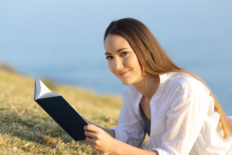 Satisfied woman reading a book looking at camera lying on the grass. Satisfied woman reading a book looking at camera lying on the grass