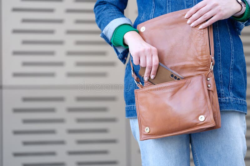 Woman putting mobile phone in brown leather handbag closeup. Storage and organization concept. Woman putting mobile phone in brown leather handbag closeup. Storage and organization concept